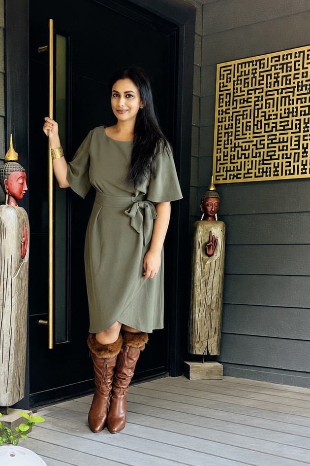 Anabin in an olive wrap dress and tan fur-trimmed boots at her black front door, between two carved Buddha figures