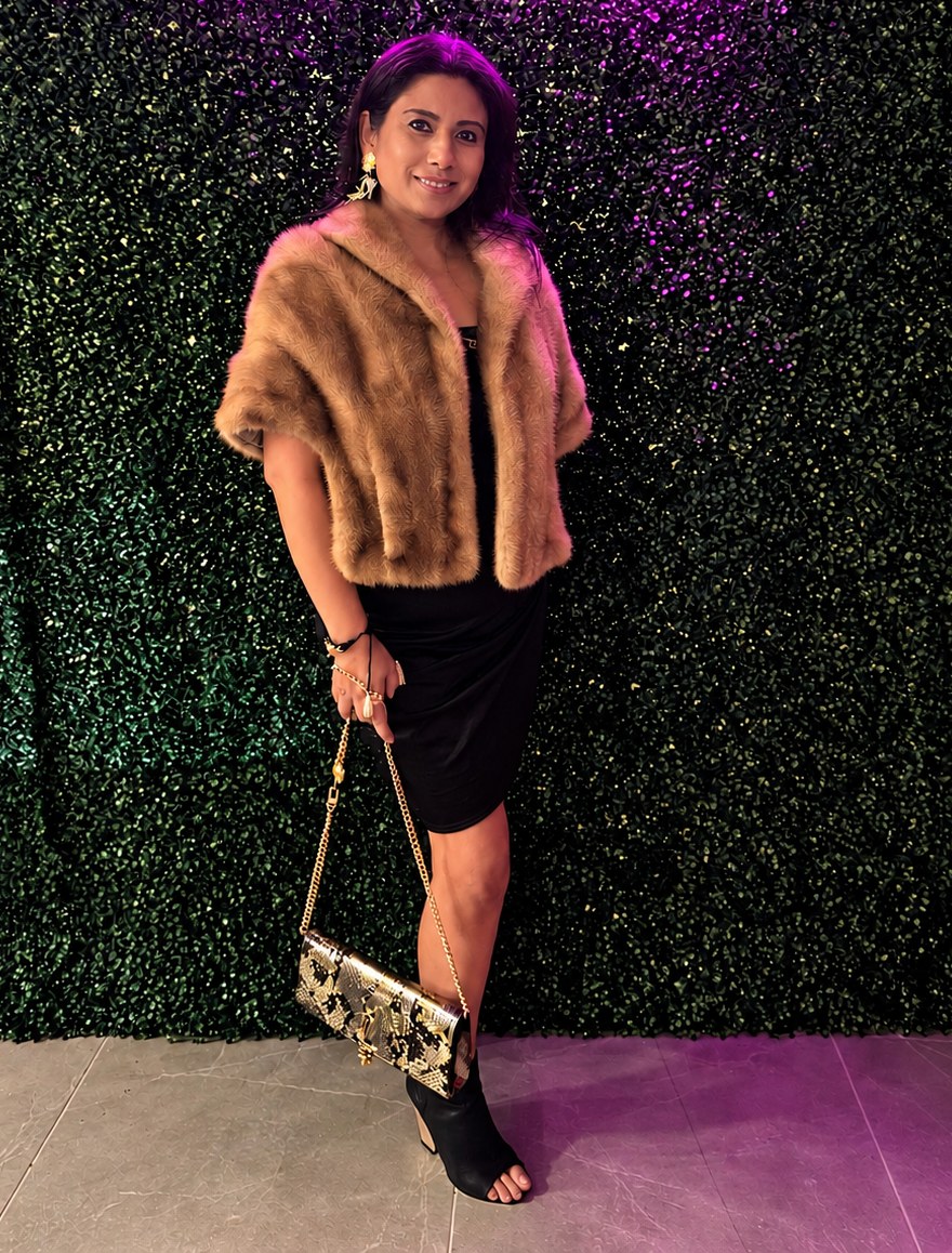 Founder in a mink stole and black dress with a gold python clutch, standing against a green hedge wall